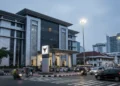 Tiga pria di Makassar ditangkap karena promosi judi online yang menghasilkan laba sebesar Rp 1,3 Miliar. Latar belakang berupa bangunan Polres Makassar dengan desain modern dan cahaya lampu jalan yang
