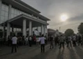 Sebuah adegan protes di luar Mapolsek Makassar dengan warga yang berkumpul di depan pintu masuk. Bangunan polisi modern dengan dinding putih dan jendela besar yang menangkap cahaya sore. Pintu masuk d