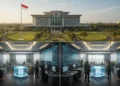 The image shows the modern headquarters of the Indonesian Armed Forces (TNI) in Jakarta, with the Indonesian national flag flying proudly. The building is surrounded by greenery and well-maintained pa