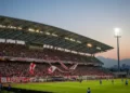 Gelora Bung Karno Stadium is filled with enthusiastic Persib fans in red and white, preparing for a match against Madura United. The modern stadium structure with its curved roof and the bustling crow
