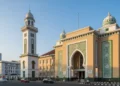 The image depicts the Makassar City Hall, a modern building with intricate Islamic architectural elements, warm beige and green accents, and a clear sky with soft sunlight. The scene captures the city
