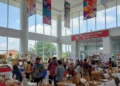 The entrance of the PKK Makassar office building is shown, with a modern atrium filled with colorful banners and stalls displaying local crafts and products. The building's exterior is clean and well-