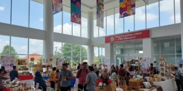 The entrance of the PKK Makassar office building is shown, with a modern atrium filled with colorful banners and stalls displaying local crafts and products. The building's exterior is clean and well-