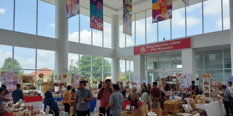 The entrance of the PKK Makassar office building is shown, with a modern atrium filled with colorful banners and stalls displaying local crafts and products. The building's exterior is clean and well-