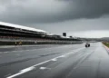 Lanskap Phillip Island Grand Prix Circuit dengan langit berawan tebal dan hujan ringan, track balap kosong dengan tarmac yang basah, grandstand dan bangunan pit terlihat dengan jelas, serta bendera Au