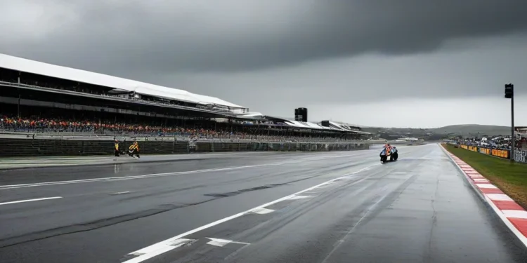 Lanskap Phillip Island Grand Prix Circuit dengan langit berawan tebal dan hujan ringan, track balap kosong dengan tarmac yang basah, grandstand dan bangunan pit terlihat dengan jelas, serta bendera Au