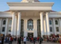 A grand entrance of Makassar City Hall with its white facade and intricate architectural details, bathed in natural light. Inside, the interior is vibrant with people setting up stalls and engaging in
