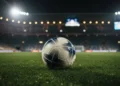 Bola sepak profesional bersandar di atas rumput stadion yang basah di bawah sorot lampu malam.