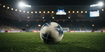 Bola sepak profesional bersandar di atas rumput stadion yang basah di bawah sorot lampu malam.