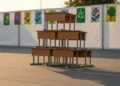 Foto close-up meja-meja kayu sekolah yang rapi disusun di area lapangan kosong saat matahari terbenam dengan cahaya hangat panjangnya bayangan dan dinding putih berposter seni pendidikan.