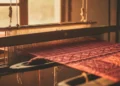 "Foto detail kain sutera merah dengan motif anyaman halus di atas alat tenun kayu, dipancing cahaya matahari sore yang hangat sebagai metafora ikatan sosial masyarakat Bugis"