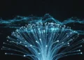 Foto makro kabel serat optik bersinar yang saling bertaut dengan aliran data transparan berbentuk hologram melayang di atasnya terhadap latar belakang hitam pekat. Cahaya biru volumenya menerangi pola