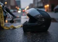 Alhamdulillah, Malam Ini Mulai Tarawih 1 Ramadan 1444H Foto close-up helm motor dan tali polisi kuning di aspal basah dengan cahaya kota buram.