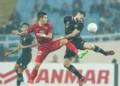 Putaran Kedua MILO Football Championship di Makassar Jersey emas dengan hiasan benang bordir detail bertekstur diletakkan di atas meja kayu berkilau, diterangi lampu sorot stadion dramatis pada malam hari.