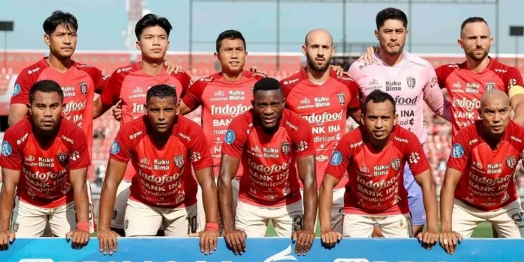 Bola sepak profesional bersandar pada rumput basah bawah sorot lampu stadion dramatis malam hari dengan fokus tajam tekstur detail menunjukkan tetesan air yang memantulkan cahaya sekitar tas peralatan