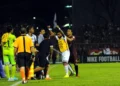 Suasana pertandingan sepak bola malam hari dengan hujan deras dan sorot lampu stadion yang dramatis memotong kabut. Sebuah bola sepak putih sempurna berada di atas rumput basah dekat sepatu boot profe