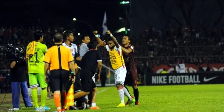 765 Personel Gabungan Amankan Laga PSM Makassar vs Madura United di Balikpapan Suasana pertandingan sepak bola malam hari dengan hujan deras dan sorot lampu stadion yang dramatis memotong kabut. Sebuah bola sepak putih sempurna berada di atas rumput basah dekat sepatu boot profe